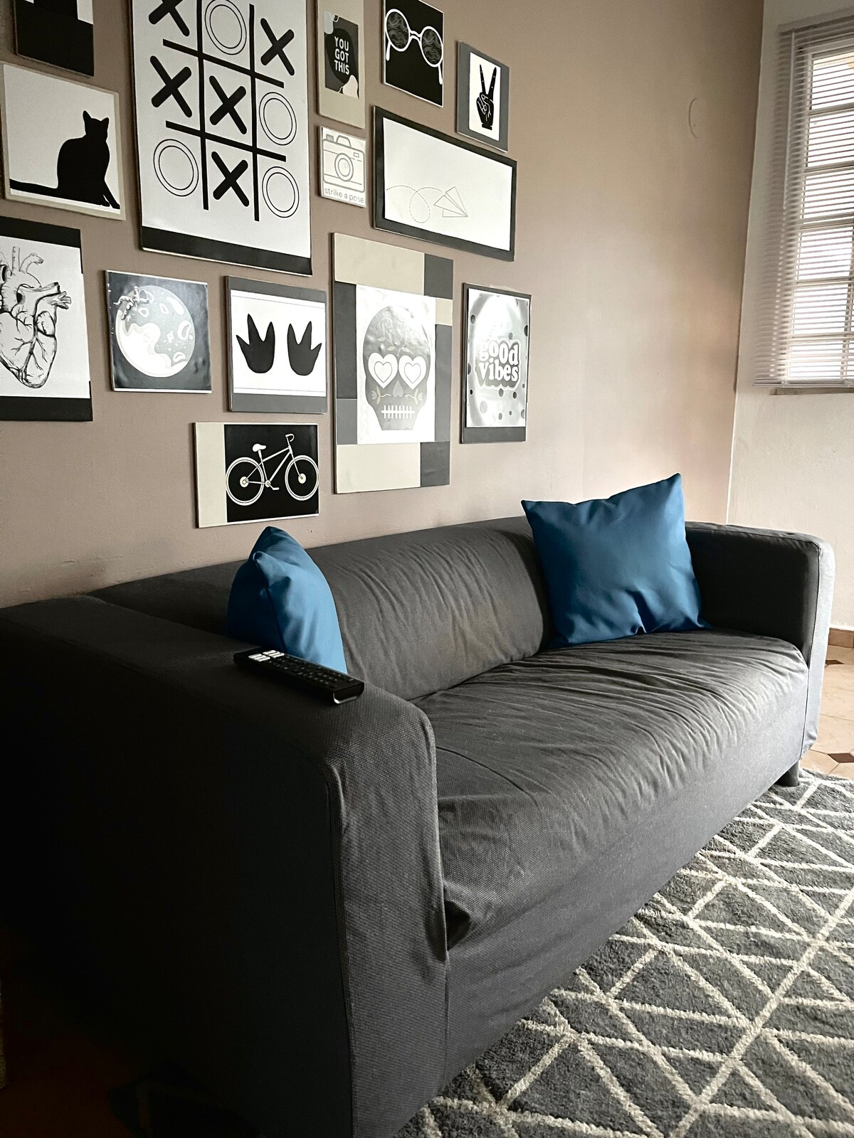 A gray sofa is positioned against a wall featuring a gallery of framed black and white artwork. Two blue cushions add a touch of color to the seating area. A remote control rests on the arm of the sofa, and natural light filters through a nearby window.