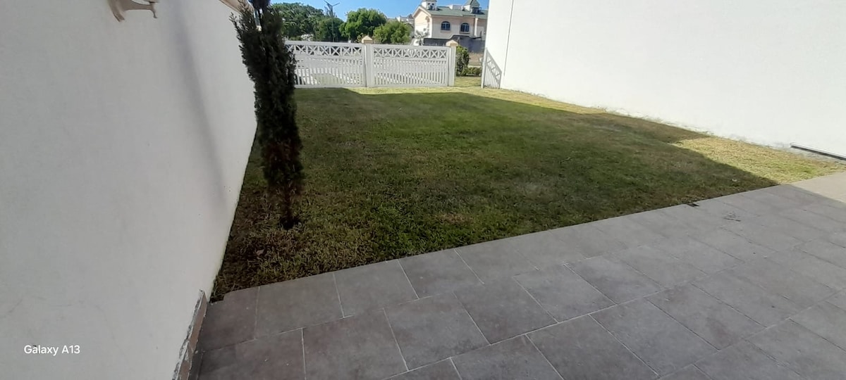 A well-maintained outdoor area features a grassy lawn bordered by a clean white wall. A patio with light-colored tiles provides a space for relaxation. The view includes nearby greenery and distant structures, contributing to a serene outdoor atmosphere.