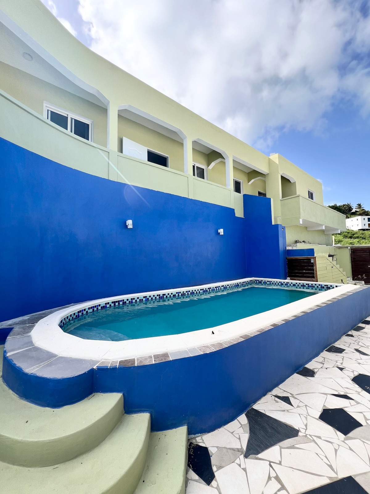 A private pool is prominently featured, surrounded by a vibrant blue wall. The pool area includes steps leading down to the water's edge, while a patterned stone surface enhances the outdoor space. The building above is painted in a soft yellow hue, contributing to a bright atmosphere.