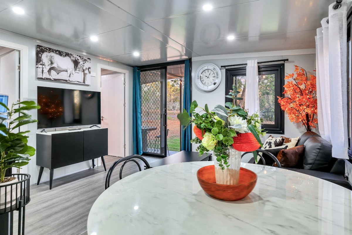 A modern living space is presented featuring a circular marble dining table with a centerpiece of fresh flowers. A large wall clock and a wall-mounted television are visible. Large windows frame the view of the outdoors, while a vibrant tree adds a touch of color.