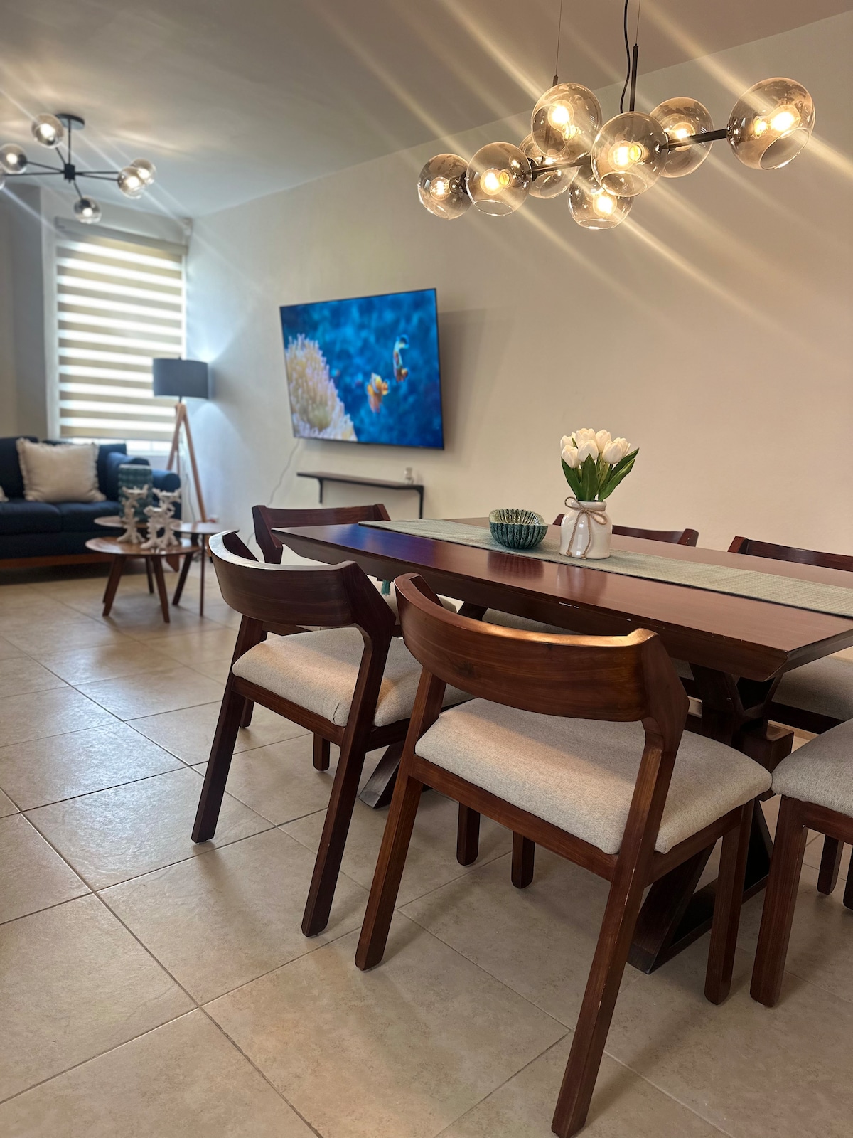 A dining area features a long wooden table surrounded by several seated chairs. A modern light fixture hangs above, providing soft illumination. A wall-mounted television displays an aquatic scene, while a vase with white flowers decorates the table. A cozy seating area is visible in the background.