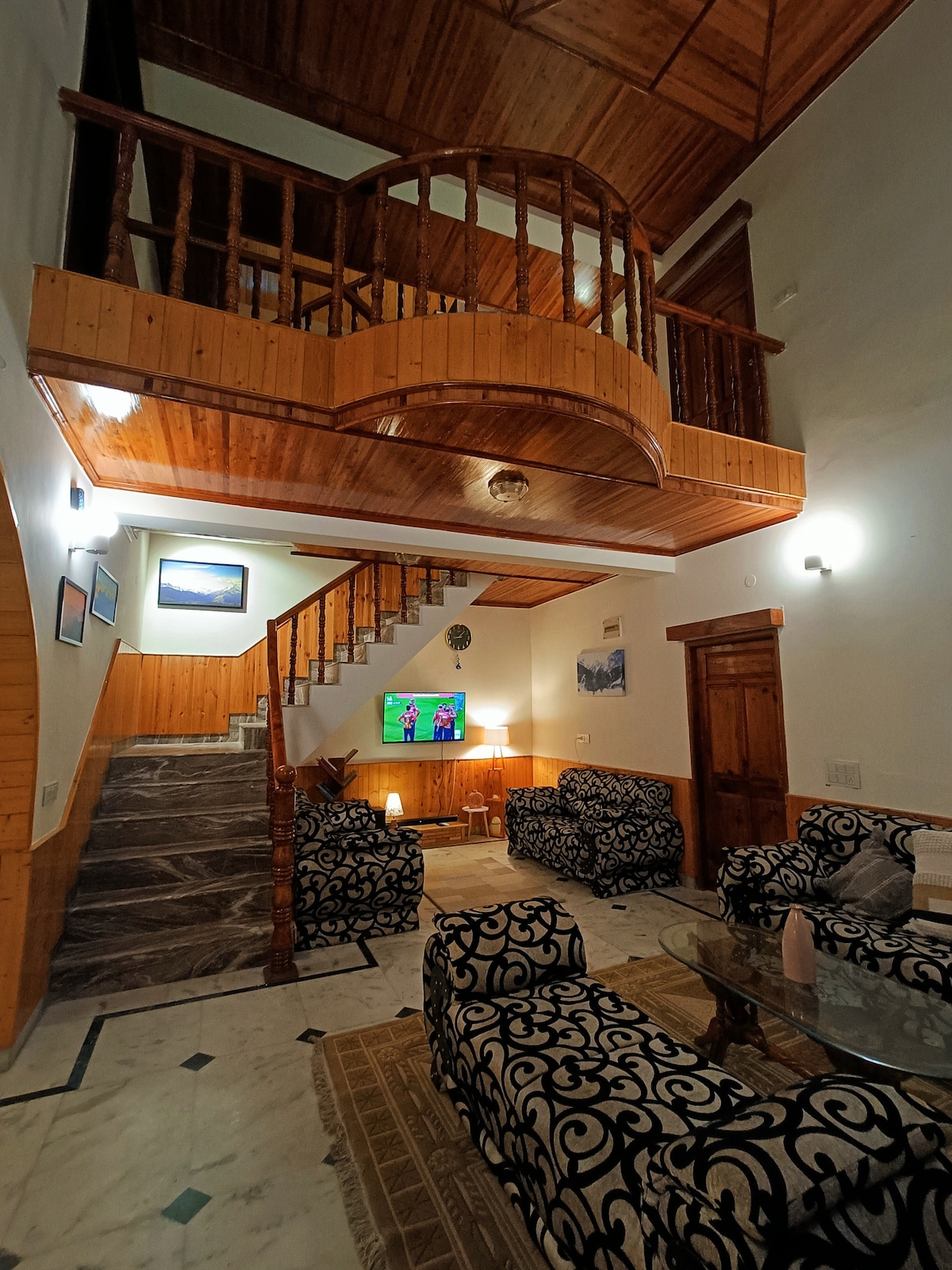 A multi-level living area features a wooden balcony overlooking a spacious seating arrangement. Two patterned sofas are positioned around a glass coffee table, with a television mounted on the wall. Natural light from nearby windows highlights the marble flooring and wooden accents.