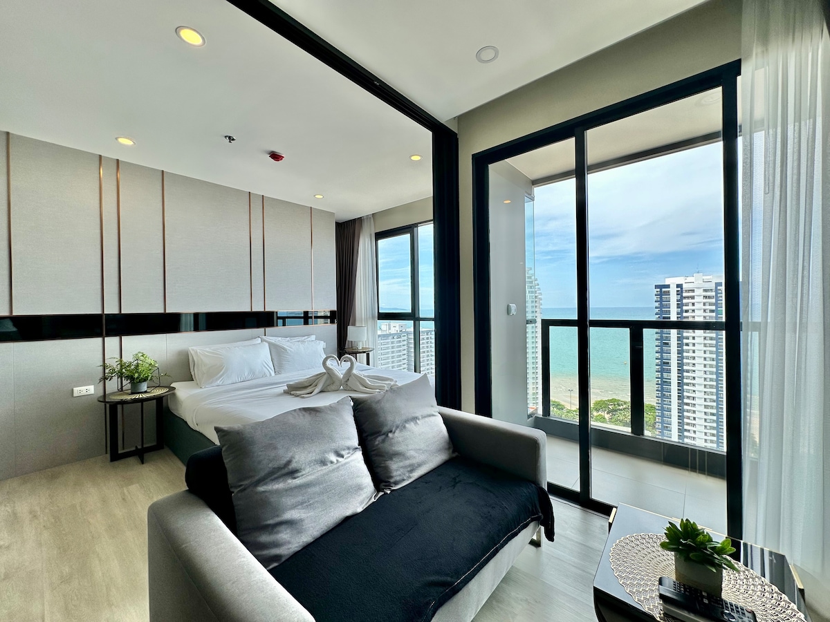A spacious studio apartment features a king-size bed with soft linens, a cozy seating area with decorative cushions, and large windows offering sea views. Natural light floods the room, enhancing the modern design elements and clean lines throughout. A small balcony provides additional outdoor access.