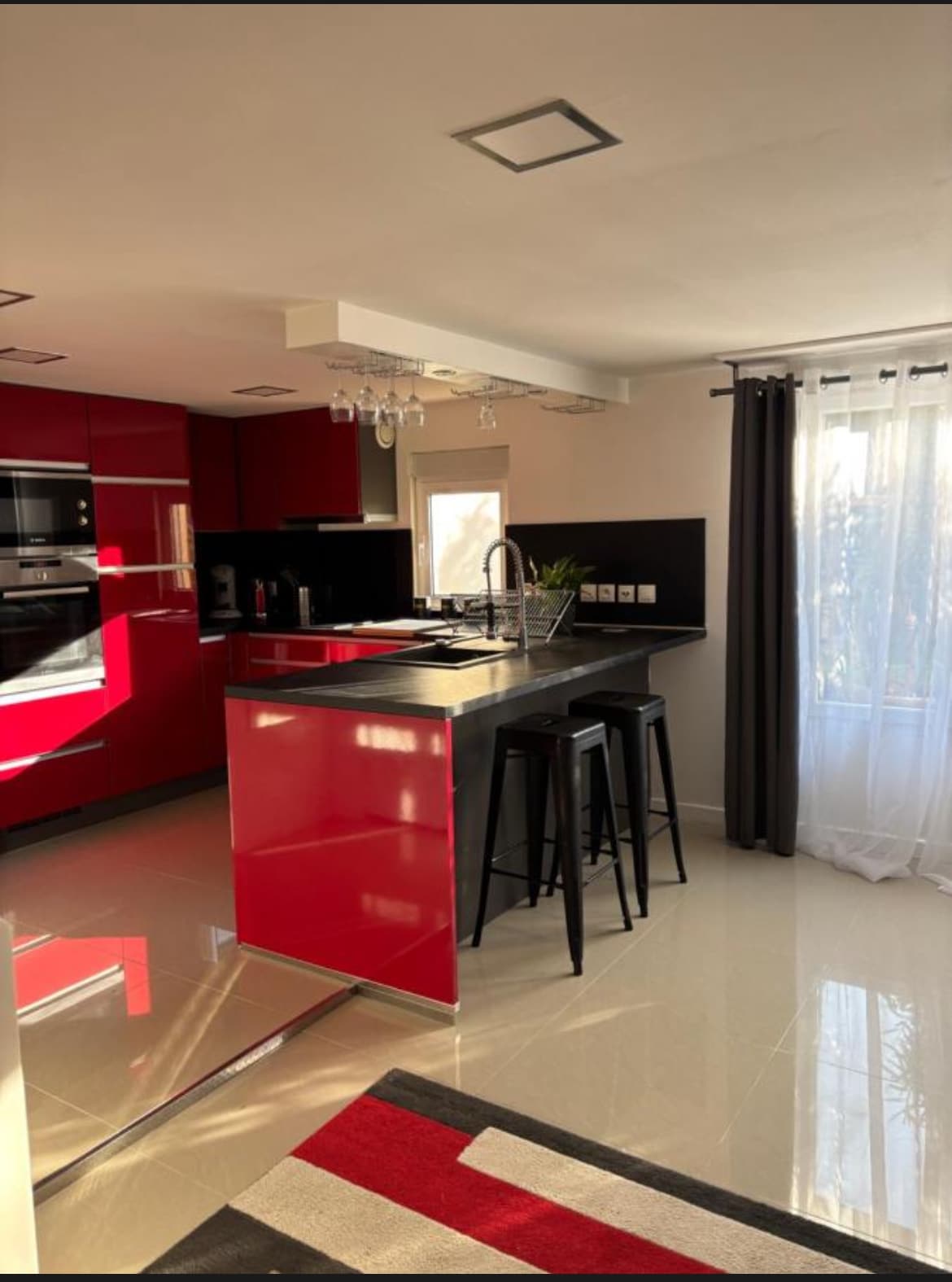 A modern kitchen features sleek red cabinetry and a central island with seating for two. Stainless steel appliances and dark countertops contribute to the kitchen's functionality. Natural light filters through a nearby window adorned with light curtains, illuminating the glossy tiled floor.