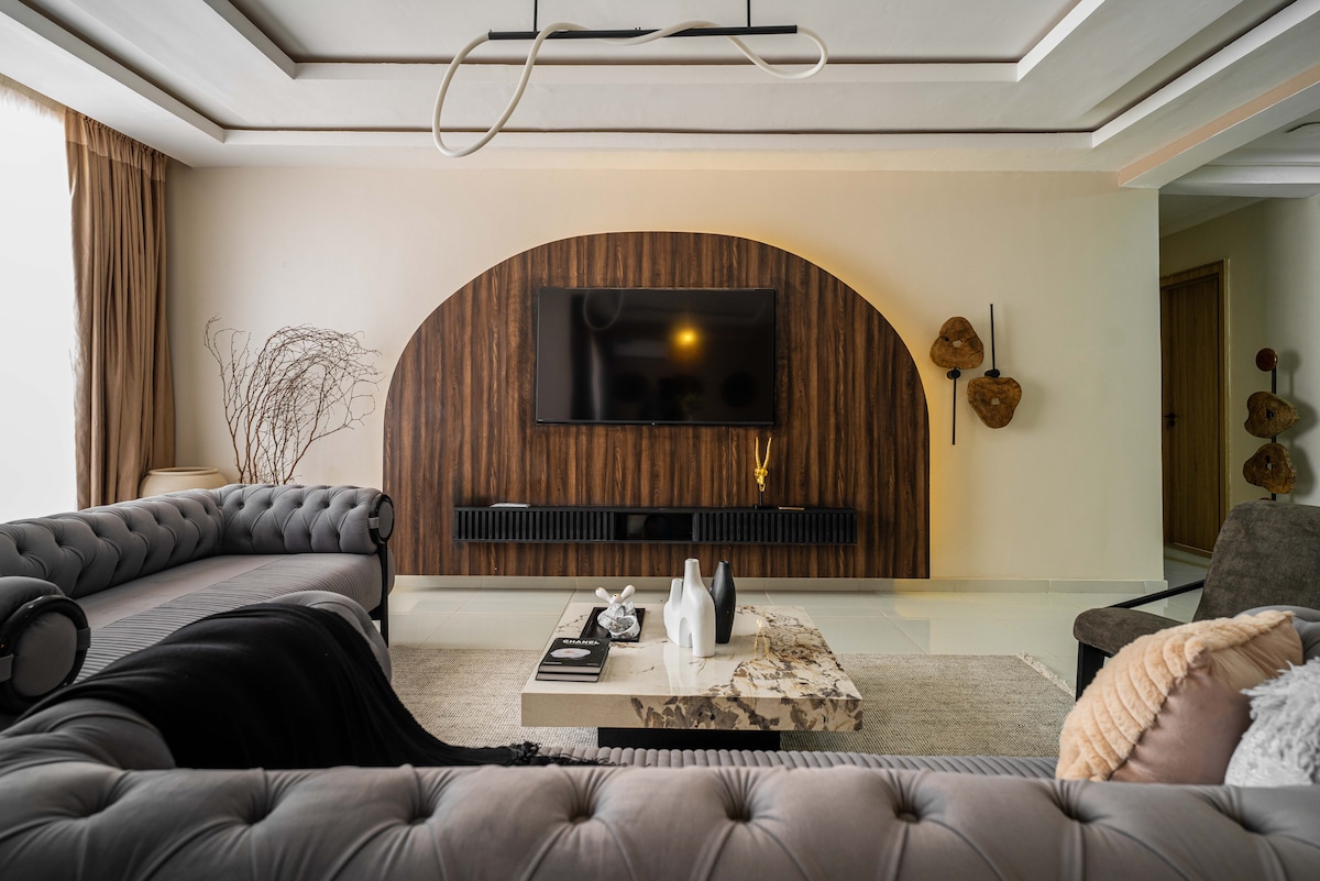 A modern living area features a wooden accent wall with a mounted television. Two plush sofas flank a central coffee table adorned with decor items. Natural light filters in through large windows, illuminating the soft rug that anchors the space.