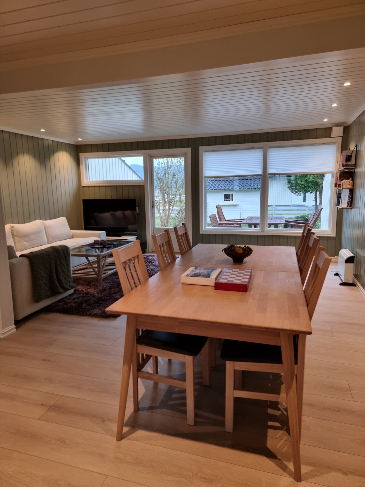A spacious living and dining area features large windows that allow natural light to fill the space. A wooden dining table with chairs is positioned adjacent to a comfortable seating area, which includes a couch and a TV mounted on the wall.
