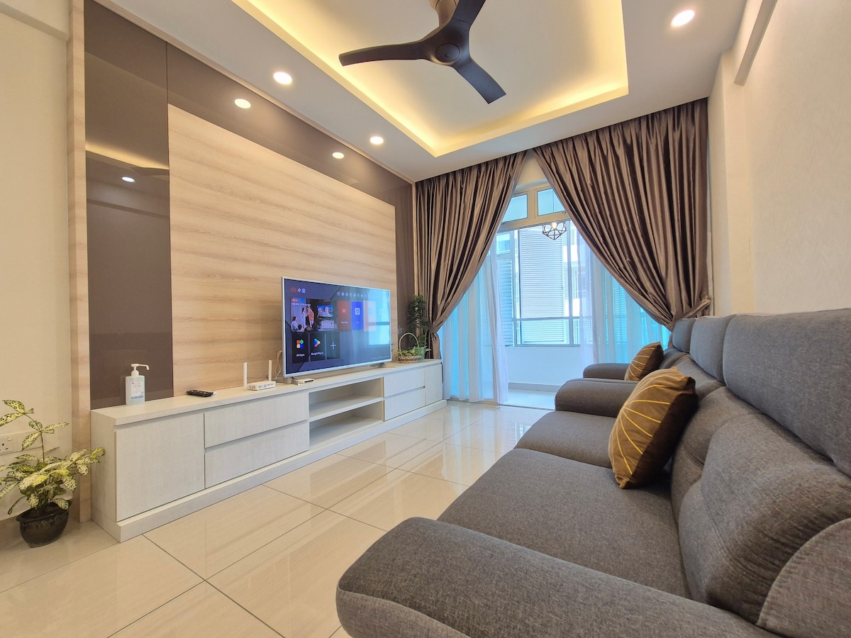 A modern living area features a sleek entertainment unit with a flat-screen TV, complemented by soft grey sofas adorned with decorative pillows. Natural light illuminates the space through large windows, and elegant curtains frame the view, enhancing the welcoming atmosphere of the room.
