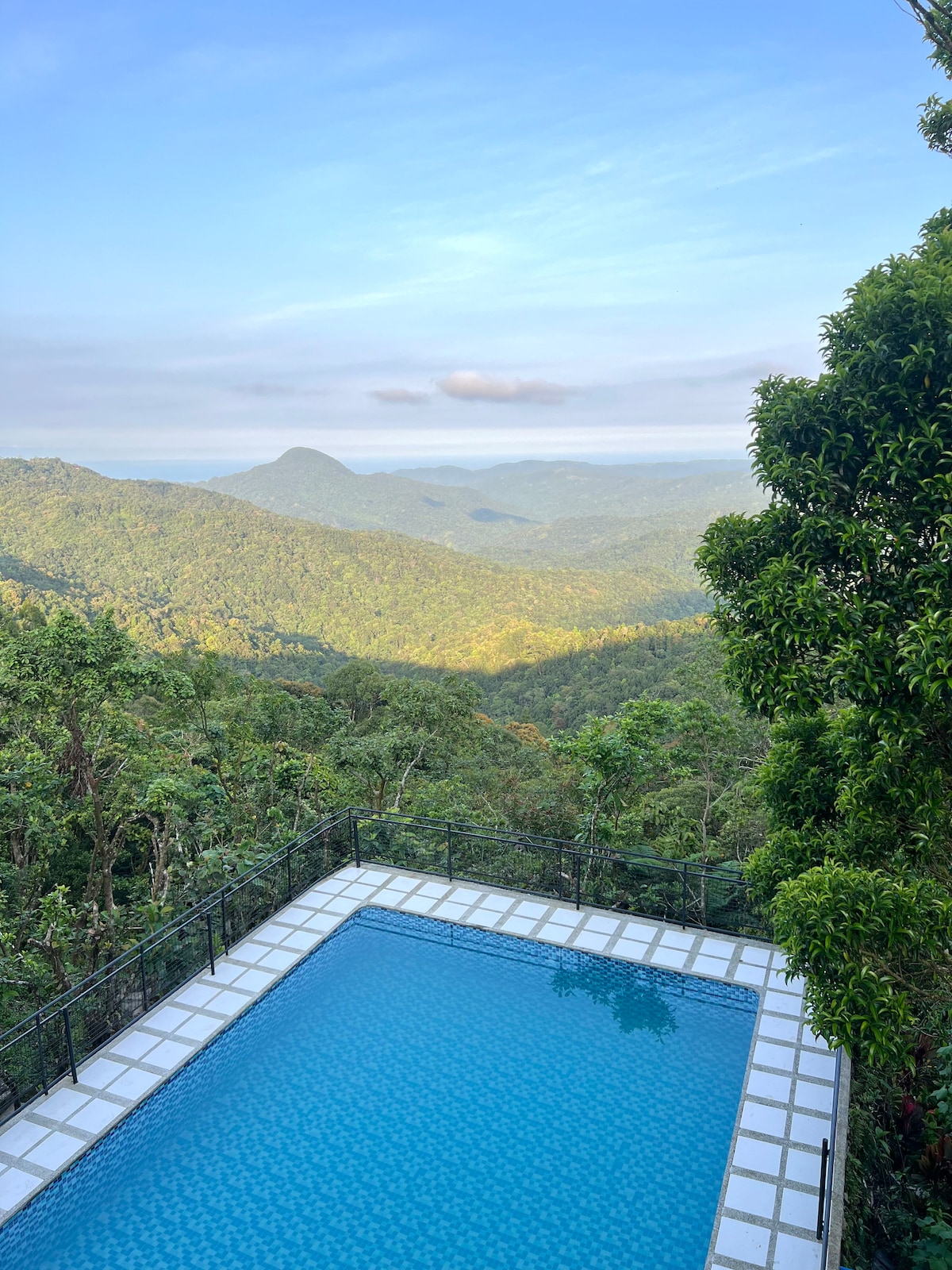 A serene outdoor swimming pool glistens in the sunlight, surrounded by a scenic mountain landscape. Lush greenery fills the view, showcasing rolling hills and valleys under a clear blue sky, inviting relaxation and connection to nature.