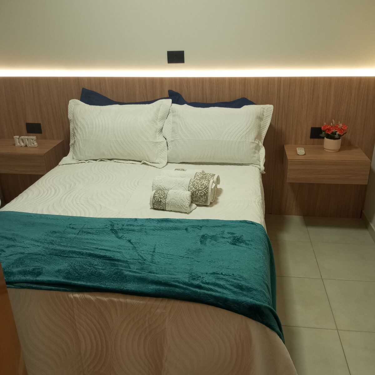 A cozy sleeping area features a spacious bed with white bedding and decorative throw pillows. Nightstands flank the bed, accentuated by soft lighting mounted above. A neatly arranged set of towels is placed at the foot of the bed, creating an inviting space.