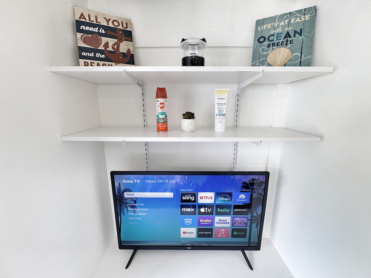 A well-organized entertainment area features a Roku TV prominently displayed on a low shelf. Above, two white shelves hold decorative items, including framed quotes, a lantern, and personal care products, alongside a small potted plant. The overall arrangement provides functionality and aesthetic appeal.