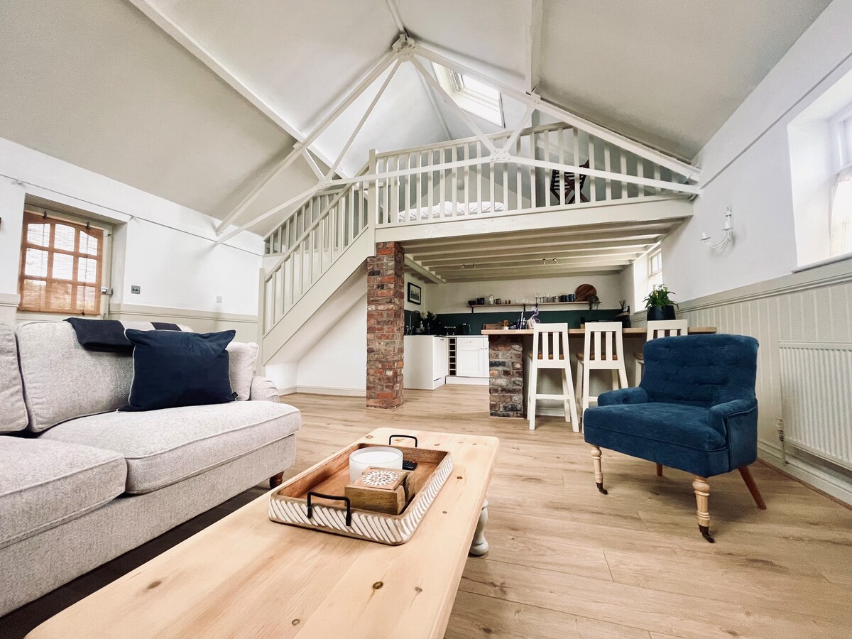 The interior space showcases a high, vaulted ceiling with wooden beams. A light-colored sofa and a blue chair are positioned near a wooden coffee table. The open-plan layout features a dining area with white chairs and a view of the kitchen in the background.