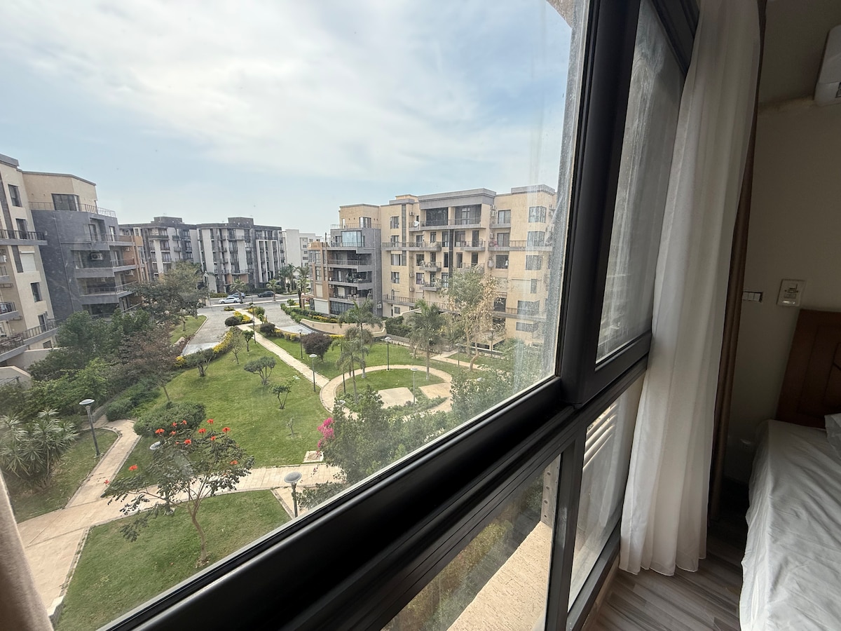 A view from a window showcases a manicured green lawn surrounded by buildings. Flowering plants add color to the landscape, while pathways meander through the area. Natural light illuminates the interior, highlighting the room's relaxed atmosphere.