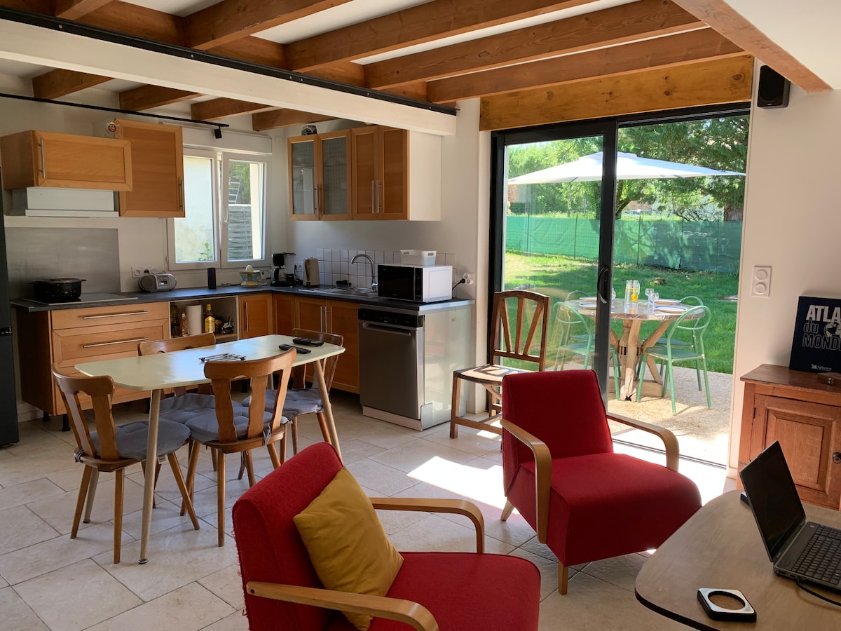 A bright kitchen area features wooden cabinetry and modern appliances. A dining table is surrounded by four wooden chairs, while two red armchairs are positioned nearby. Large glass doors offer a view of the garden, which includes a table and chairs under an umbrella.