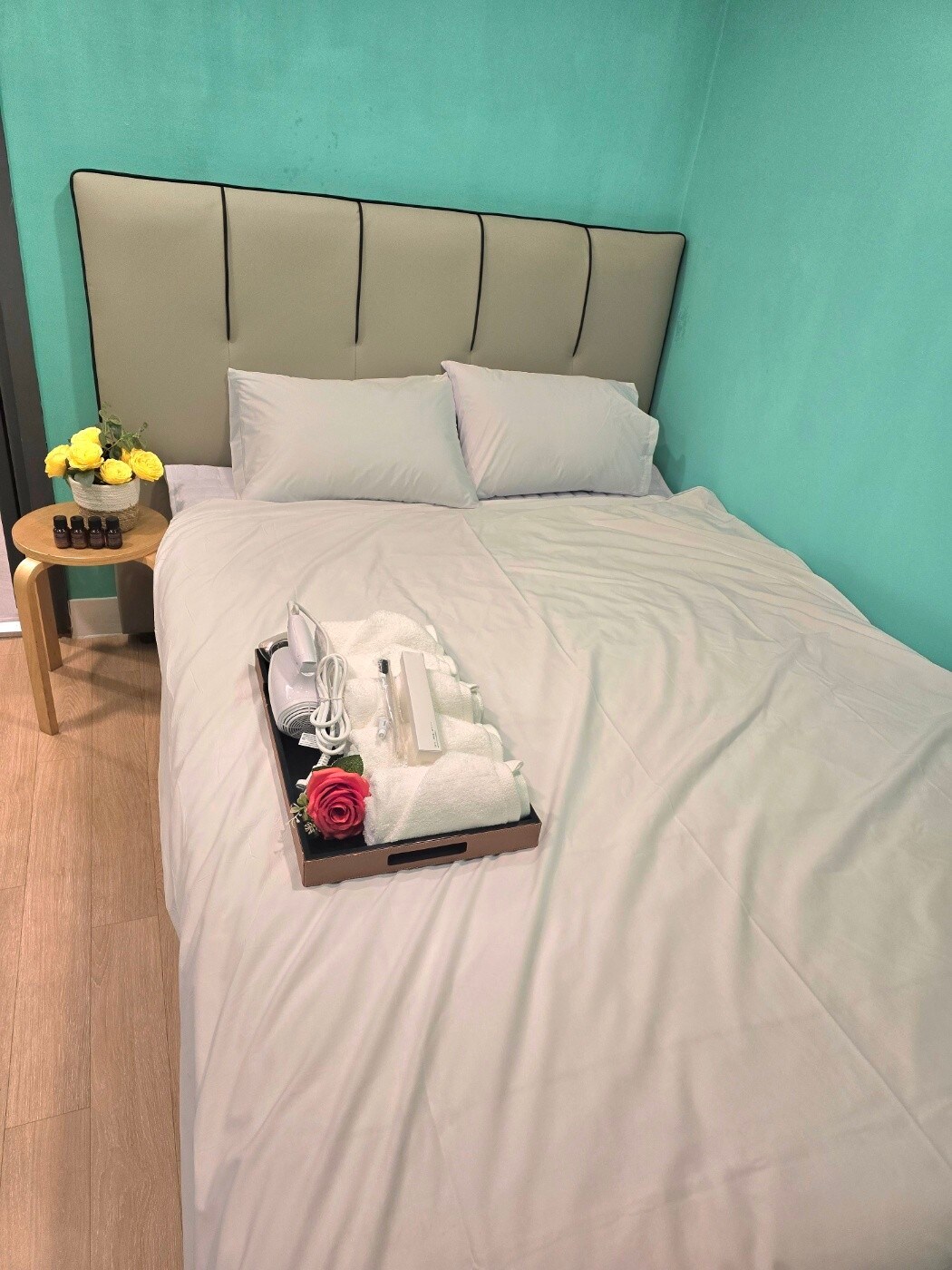 A spacious bed is positioned against a softly colored wall, adorned with fresh flowers on a side table. A tray holds neatly arranged toiletries and towels, enhancing the room's functionality and comfort.