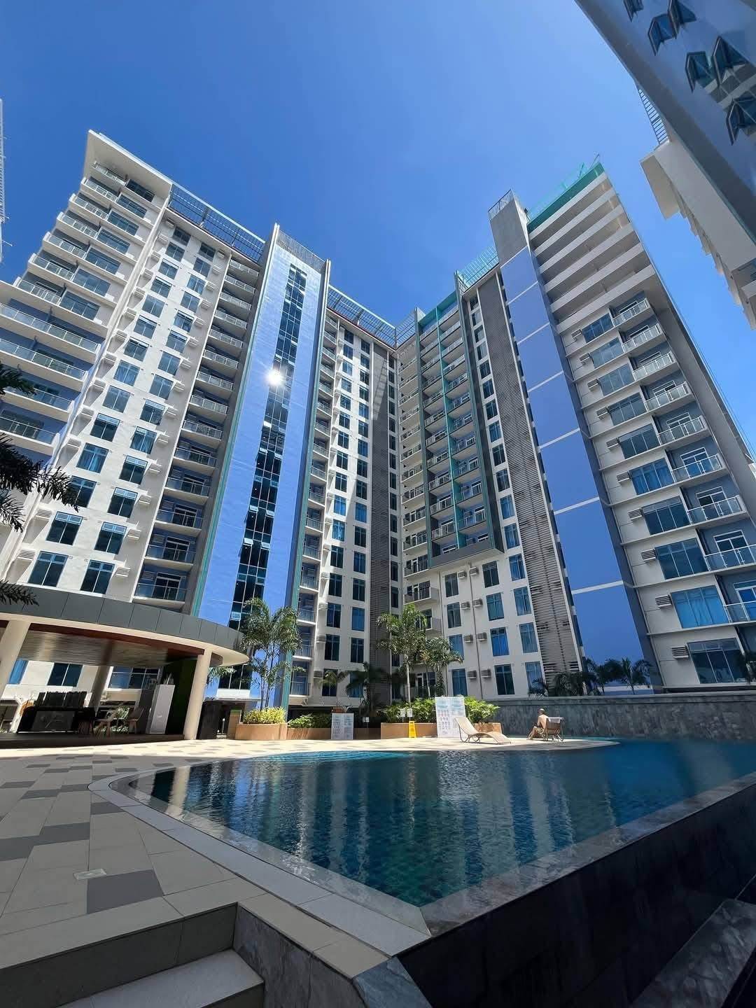 A modern high-rise building features a smooth glass façade reflecting sunlight. Surrounding the inviting pool, palm trees provide a touch of greenery. A shaded seating area is visible nearby, alongside pathways leading to the poolside.