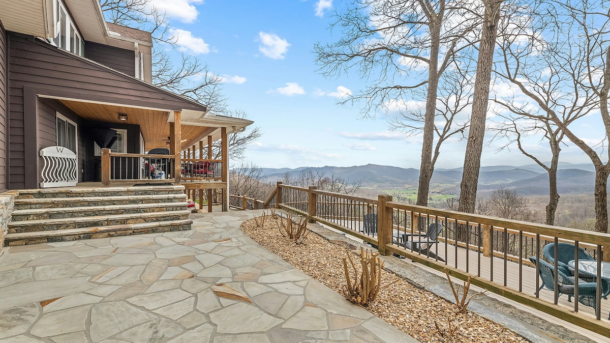 A spacious patio features a stone surface with wooden railings, providing expansive views of rolling hills and tree-lined landscapes. Comfortable seating arrangements are visible, creating an inviting space for gatherings. The surrounding area includes natural landscaping with bare trees and carefully arranged mulch.