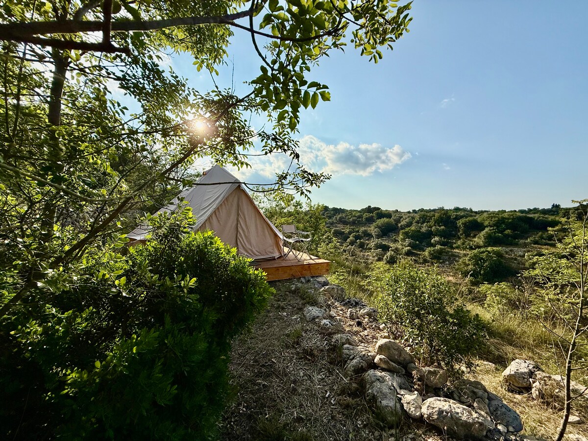 A glamping tent is nestled among greenery, surrounded by lush vegetation under a clear blue sky. Sunlight filters through the trees, highlighting the tent's fabric exterior. The surrounding landscape features rolling hills, creating a serene and spacious atmosphere conducive to nature exploration.