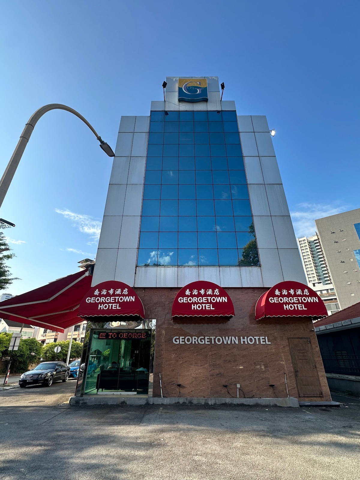 The exterior of Georgetown Hotel is showcased, featuring a modern, glass-fronted structure with large windows reflecting the sky. A covered entrance with red awnings displaying the hotel name adds a welcoming touch. Surrounding elements include a street and neighboring buildings.