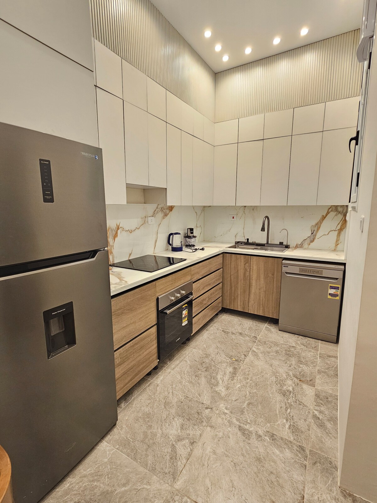 A modern kitchen features sleek cabinetry in light tones, complemented by a marble countertop. A stainless steel refrigerator and dishwasher are included. The space is illuminated by recessed lighting, enhancing the clean lines and functional layout suitable for meal preparation.