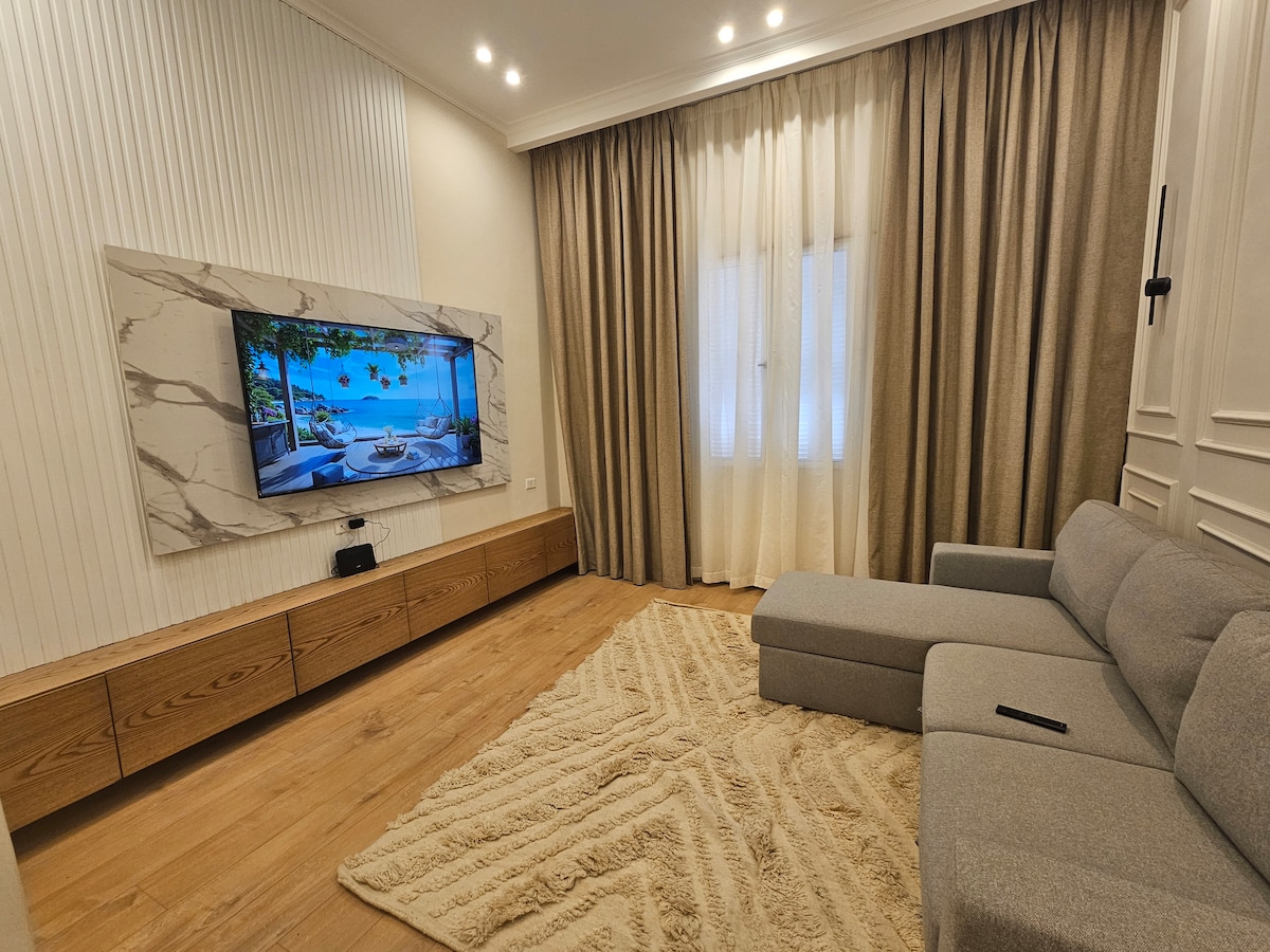 A modern living area features a plush gray sectional sofa positioned near a sleek wooden media console. A large flat-screen TV is mounted above, displaying a serene image. Soft, textured rug covers the wooden floor, and sheer curtains frame a nearby window, allowing natural light to filter in.