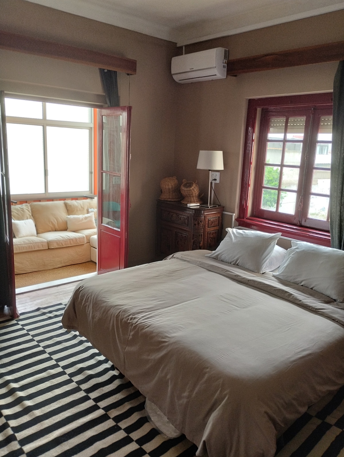 A bedroom features a spacious bed dressed in neutral linens, situated on a striped area rug. A small seating area with a light-colored sofa is visible through glass doors, which allow natural light to enter the room. An air conditioning unit is mounted on the wall.