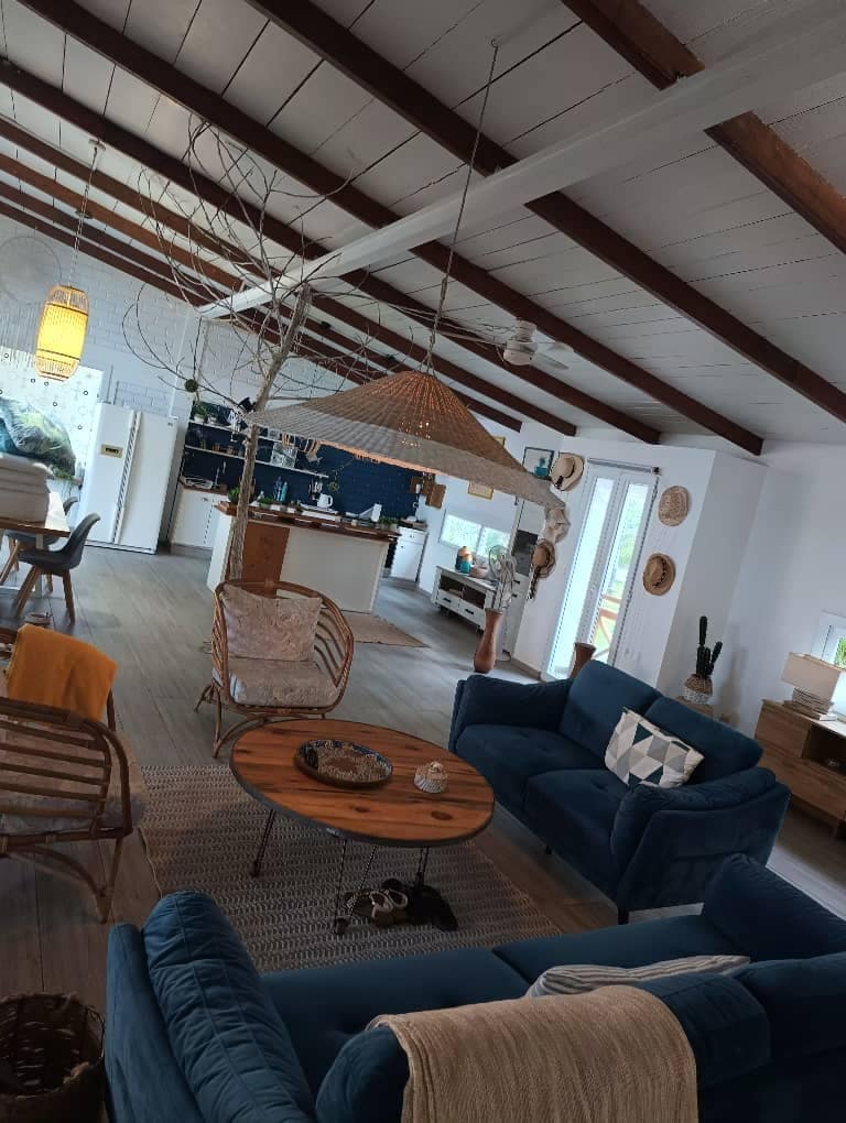 The living area features comfortable blue seating and a circular wooden coffee table, adorned with decorative items. A hanging light fixture illuminates the space, while natural light enters through large windows. The open layout seamlessly connects to the kitchen area, visible in the background.