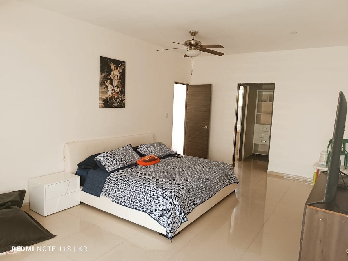 The spacious bedroom includes a large bed with a patterned blue duvet and a subtle accent pillow. A ceiling fan hangs overhead, and light enters through an adjacent door. A minimalist nightstand and a television are positioned within the room.