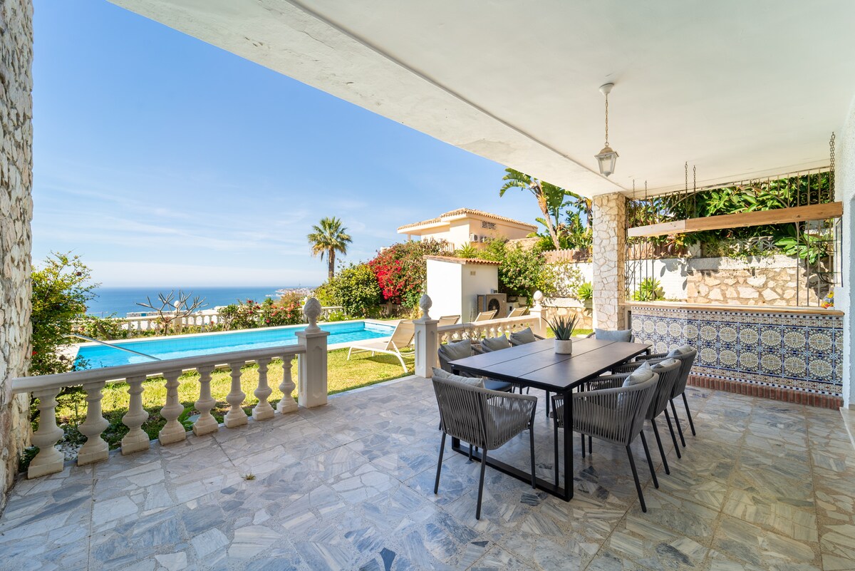 An outdoor dining area is shaded from the sun, featuring a large table surrounded by several chairs. A swimming pool, framed by manicured gardens, is visible in the background, alongside a view of the sea. Colorful flowers and greenery add to the tranquil ambiance.