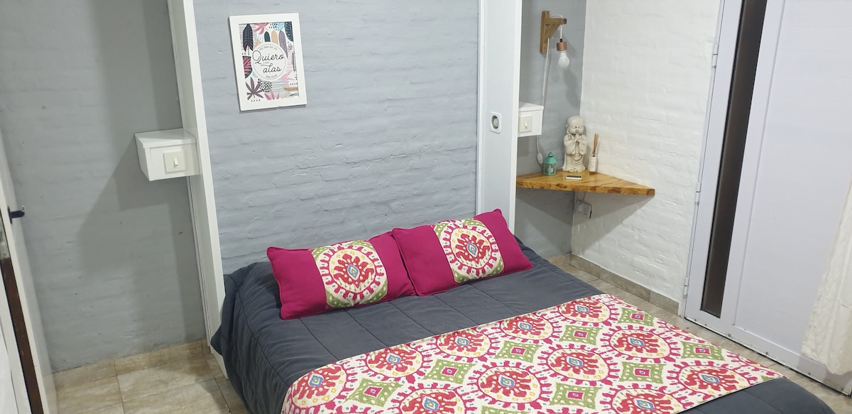 A cozy sleeping area is featured, with a bed adorned in colorful patterned bedding. Two vibrant pillows are placed against a gray wall. A small wooden shelf is positioned above the bed, displaying decorative items, while soft lighting is provided by a hanging fixture.