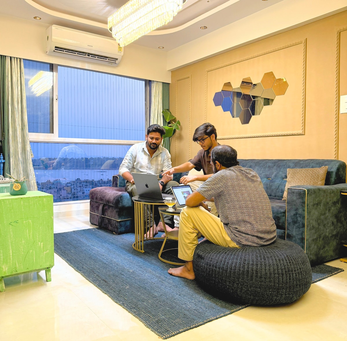 A modern living area featuring a plush sofa and a round ottoman. Large windows provide a view of the Mumbai skyline. A coffee table holds laptops while soft lighting enhances the contemporary decor. Textured walls complement the sleek furnishings.