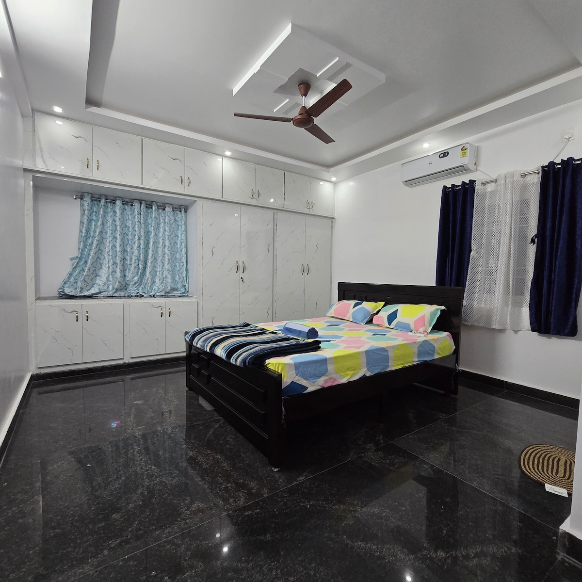 A well-lit bedroom features a spacious bed with a colorful quilt. Floor-to-ceiling cupboards provide ample storage. Blue curtains frame the window, allowing natural light to enter while maintaining privacy. A ceiling fan is positioned for air circulation in the room.