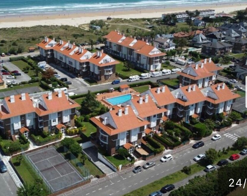 An aerial view captures a well-maintained residential complex featuring several buildings with terracotta roofs. A swimming pool is visible in the center, surrounded by landscaped gardens. Adjacent to the complex is a tennis court, and the sandy beach is seen in the background.