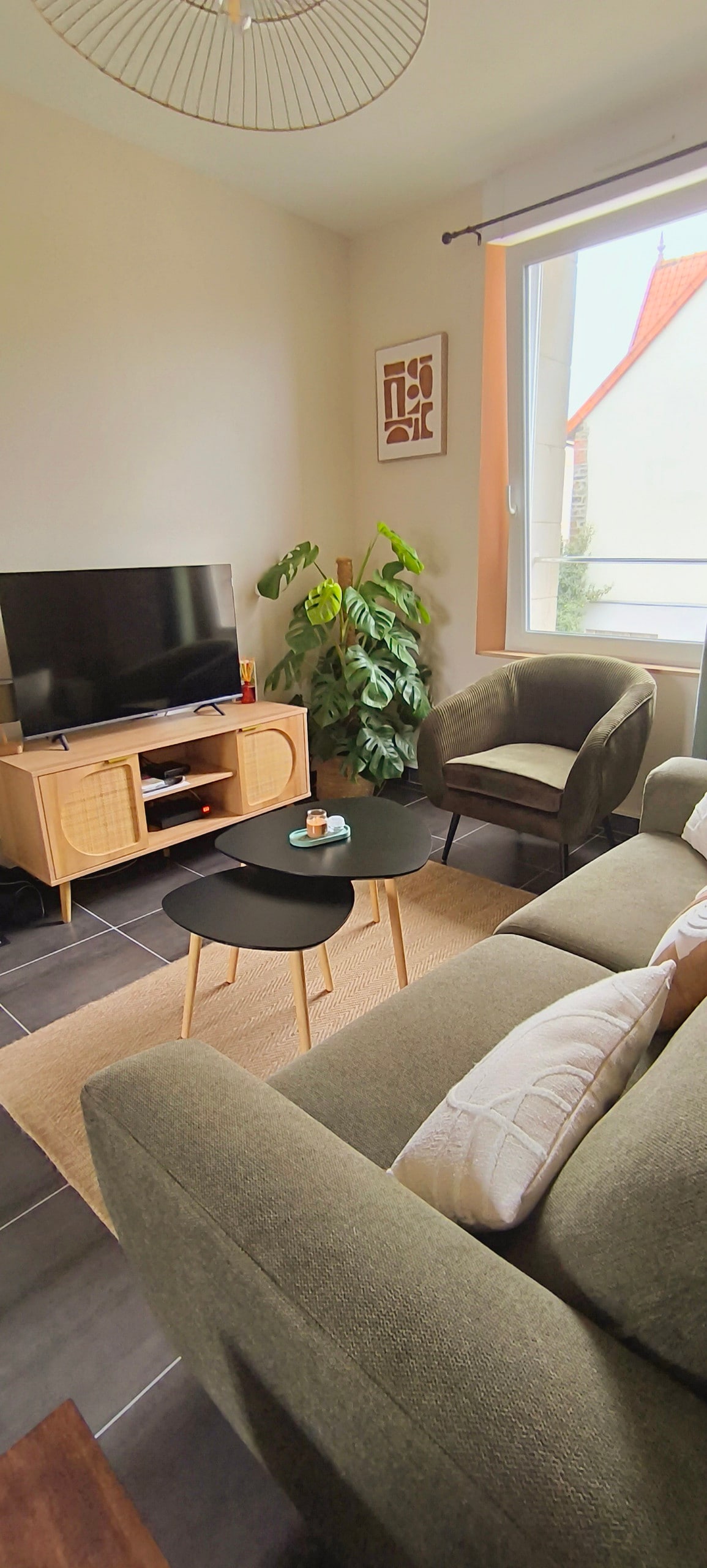 The living area is designed for comfort, featuring a modern television unit, a plush sofa, and a cozy armchair. A small black coffee table sits in front, accompanied by a potted plant, while natural light fills the room through a window with soft curtains.