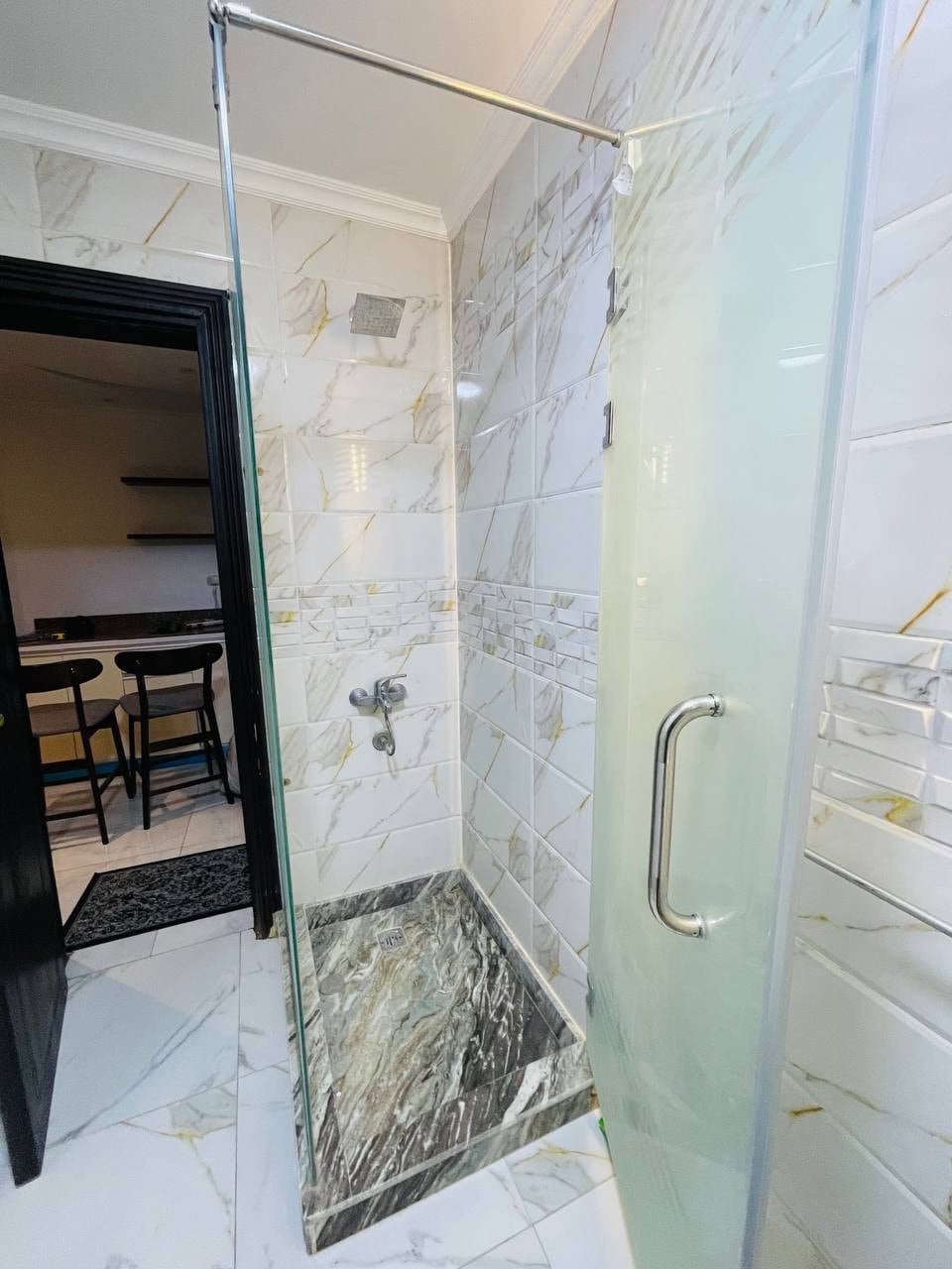 A modern shower enclosure is showcased, featuring clear glass panels and a sleek chrome handle. The walls are adorned with marble-patterned tiles, and the shower floor consists of dark, textured stone. The background hints at an open kitchen with bar seating.