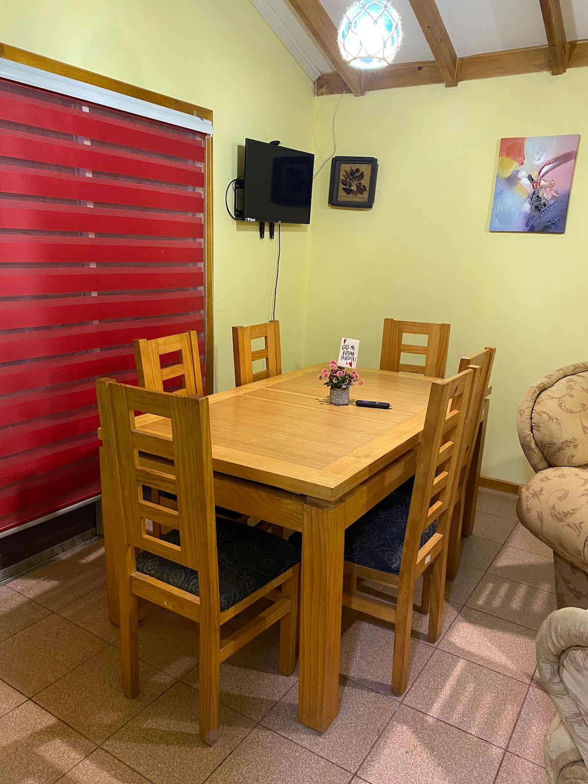 A dining area features a wooden table surrounded by six chairs. A small vase with flowers is placed at the center of the table. A wall-mounted television is visible, along with a piece of colorful artwork. Red blinds cover a window, contributing to the room's ambiance.