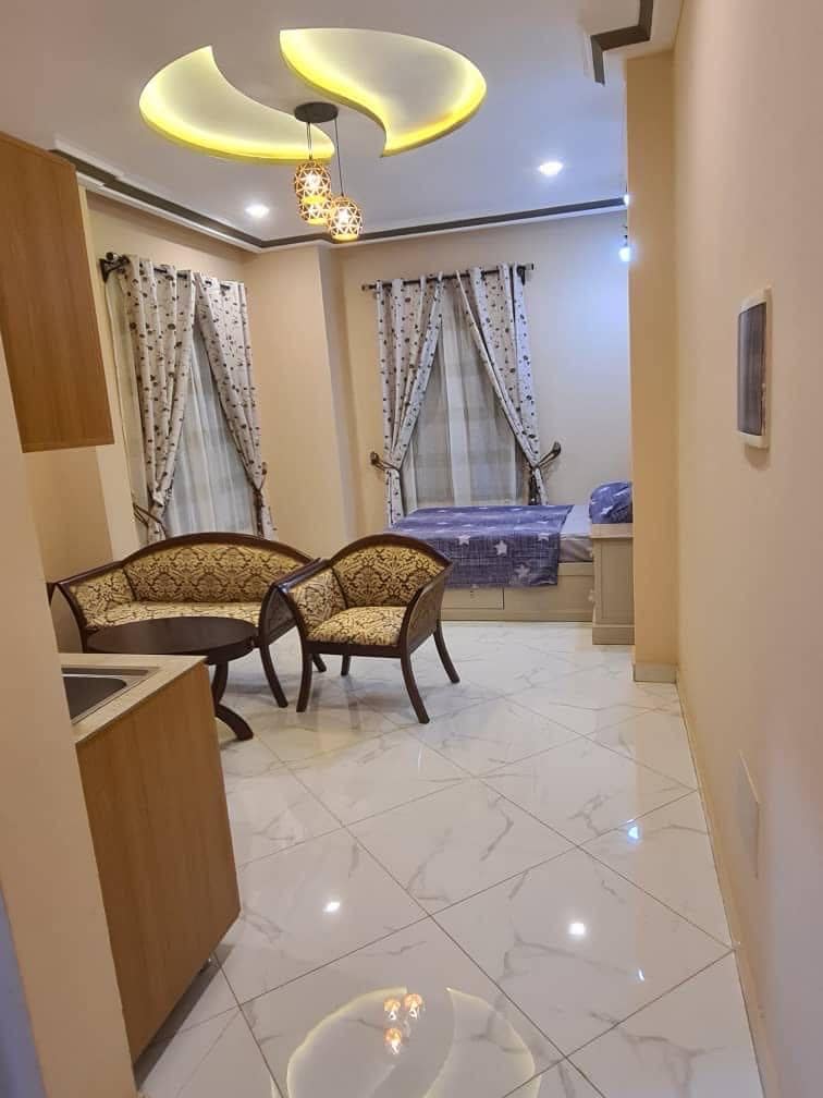 A compact living space features two elegantly designed chairs with patterned upholstery positioned adjacent to a small kitchen area. Natural light filters through sheer curtains, highlighting the glossy tile flooring. A bed with a soft cover is visible in the background, enhancing the room's cozy ambiance.
