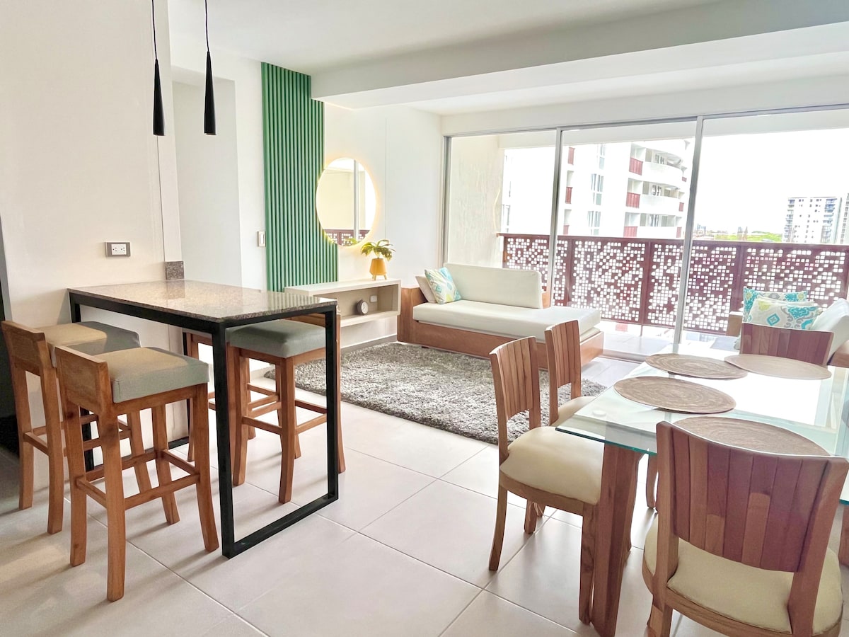 A spacious living area features large windows with natural light, creating an open atmosphere. A modern dining table with wooden chairs is seen alongside a comfortable seating arrangement, including a sofa adorned with decorative cushions. Stylish green accents enhance the contemporary decor.