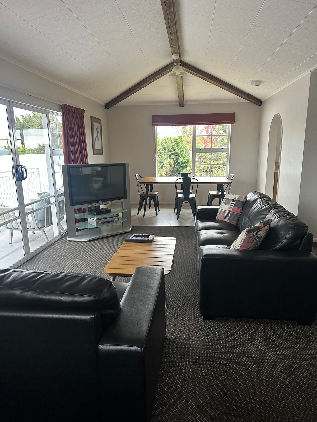 A comfortable living area is highlighted by two dark leather sofas facing a modern television. A wooden coffee table sits between them. A dining table for two is positioned near large windows, allowing for natural light and a view of greenery outside.