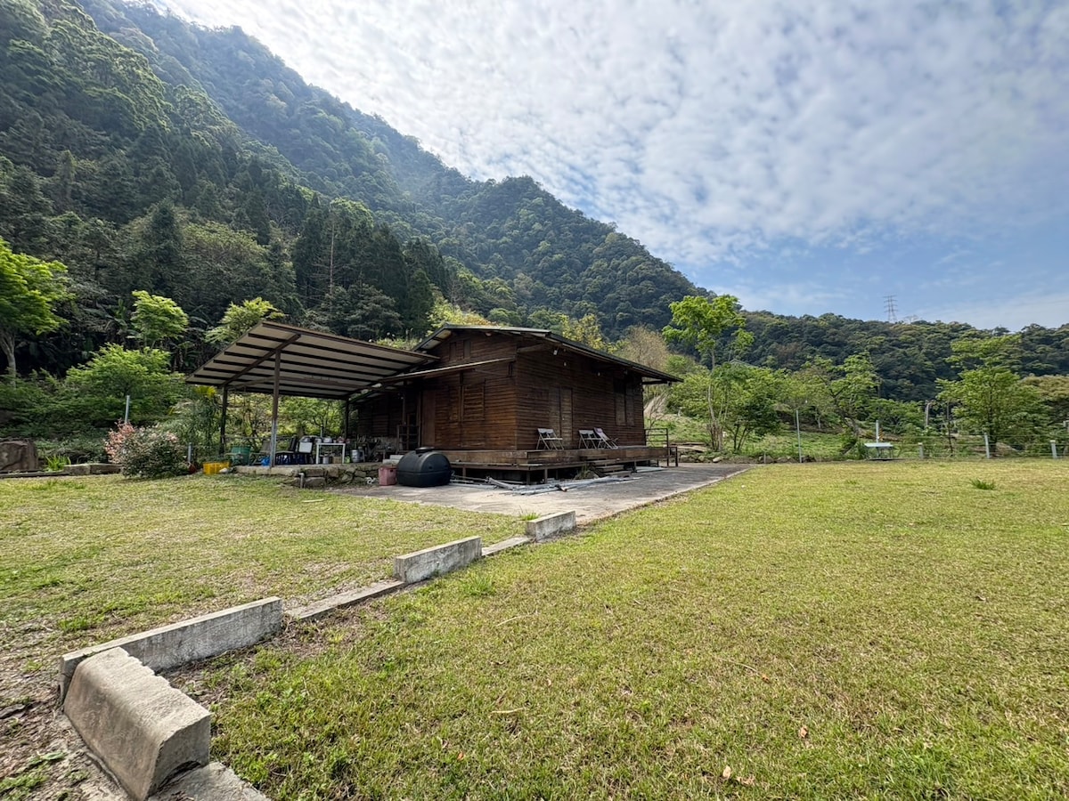 A wooden cabin is nestled in a lush green landscape, surrounded by mountains and trees. The structure features a covered outdoor area with chairs and a stage-like platform. The expansive lawn provides ample space for activities, under a sky dotted with soft clouds.