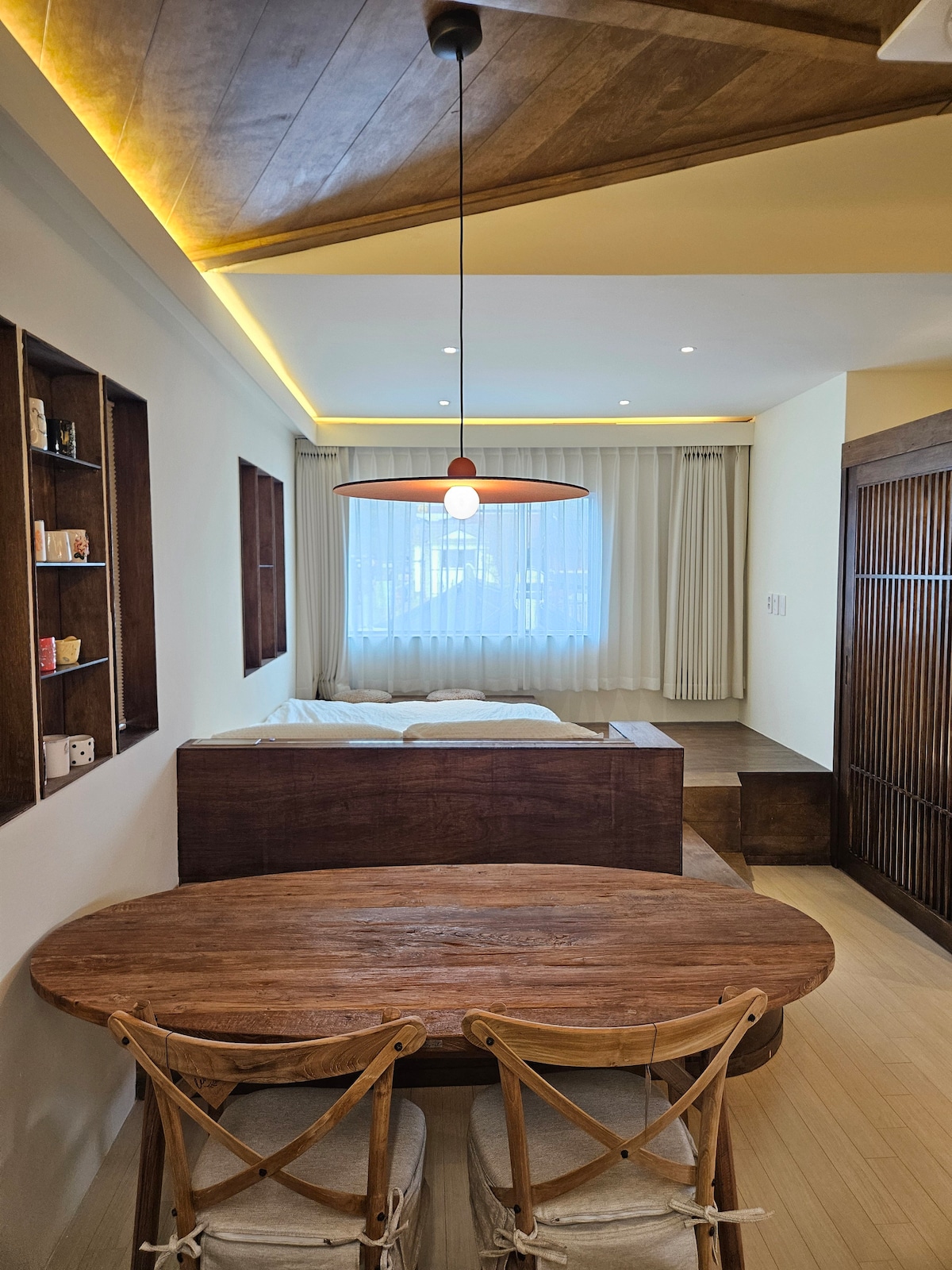 A wooden table with two chairs occupies the foreground, while a spacious bed is positioned in the background. Soft, natural light is filtered through a large window, which is dressed in sheer white curtains. Warm overhead lighting highlights the room's wooden accents.