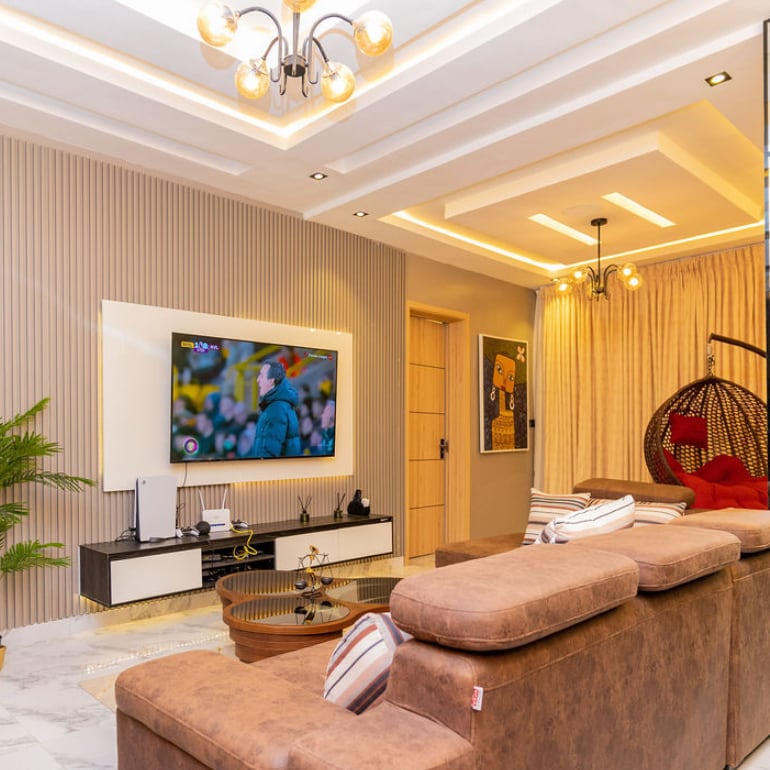 A modern living area features a comfortable brown sofa complemented by decorative pillows. A sleek television is mounted on the wall, displaying visuals. A hanging wicker chair adds an element of design, while warm lighting highlights the textured walls and elegant ceiling design.