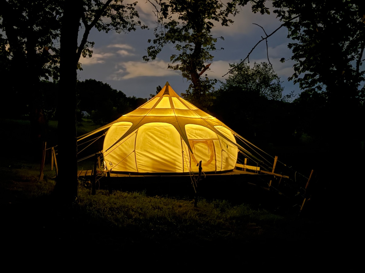 A spacious tent is illuminated in soft yellow light against a twilight backdrop, surrounded by trees. The unique structure features a pointed roof and large windows, creating a welcoming atmosphere in the midst of nature. The surrounding greenery adds to the tranquil setting.