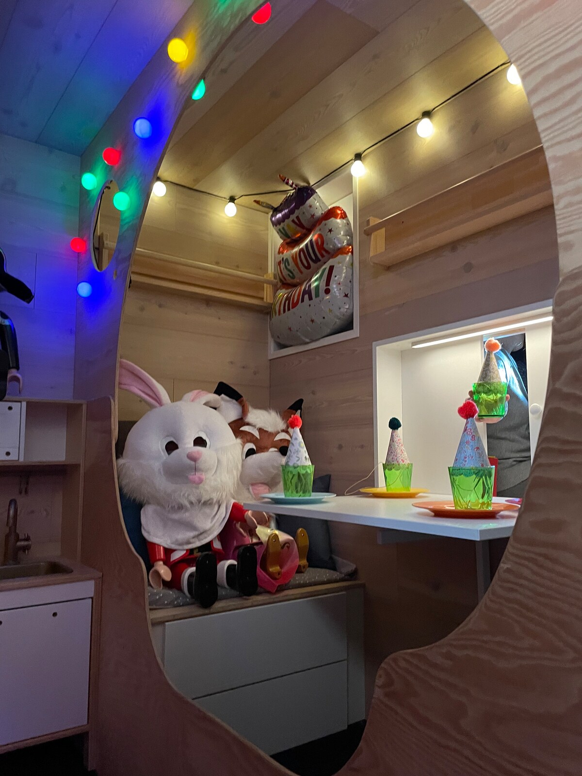 A whimsical corner of the playroom is displayed, featuring colorful party hats and plush animal costumes. Soft, twinkling lights create a cheerful ambiance, while a small table is adorned with decorative cups, enhancing the playful atmosphere perfect for children's activities.