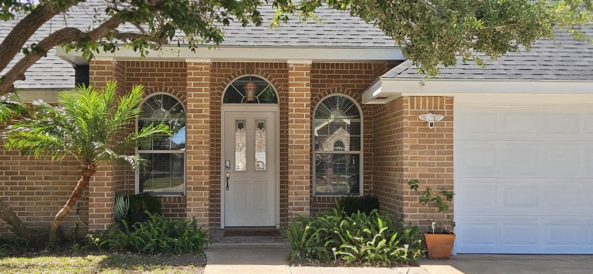 The entrance of a brick home is framed by lush greenery and palm plants. Large, arched windows let in natural light, while a covered porch offers shelter. The door, adorned with glass panels, leads into the home, which contrasts with the white garage door on the right.