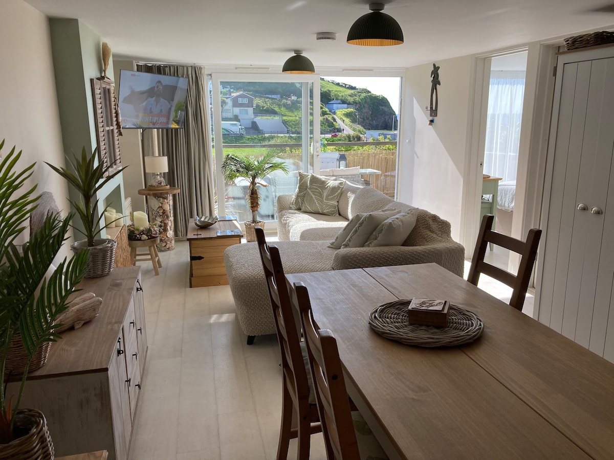 A light-filled living area features a cozy sectional sofa placed beside a large window, providing views of the surrounding landscape. A dining table is set with woven placemats, and a smart TV is mounted on the wall. Decorative elements include a potted palm and coastal-themed decor.