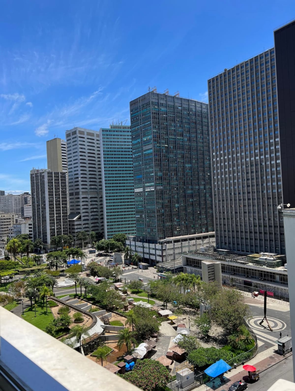 A view of the city skyline is captured from a high vantage point, featuring a mix of modern skyscrapers and greenery. Parks and open spaces are visible, along with pathways and seating areas, providing a contrast to the surrounding buildings and creating a vibrant urban atmosphere.