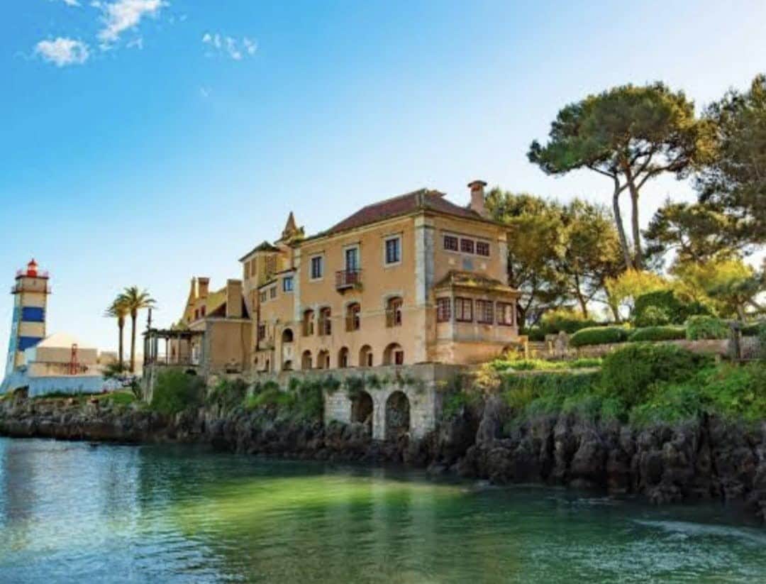 A historic building is situated along the waterfront, showcasing a blend of architectural styles. Lush greenery surrounds the property, highlighted by trees and shrubs. The calm water reflects the structure in soft hues, while a lighthouse stands in the distance, adding to the scenic coastal view.