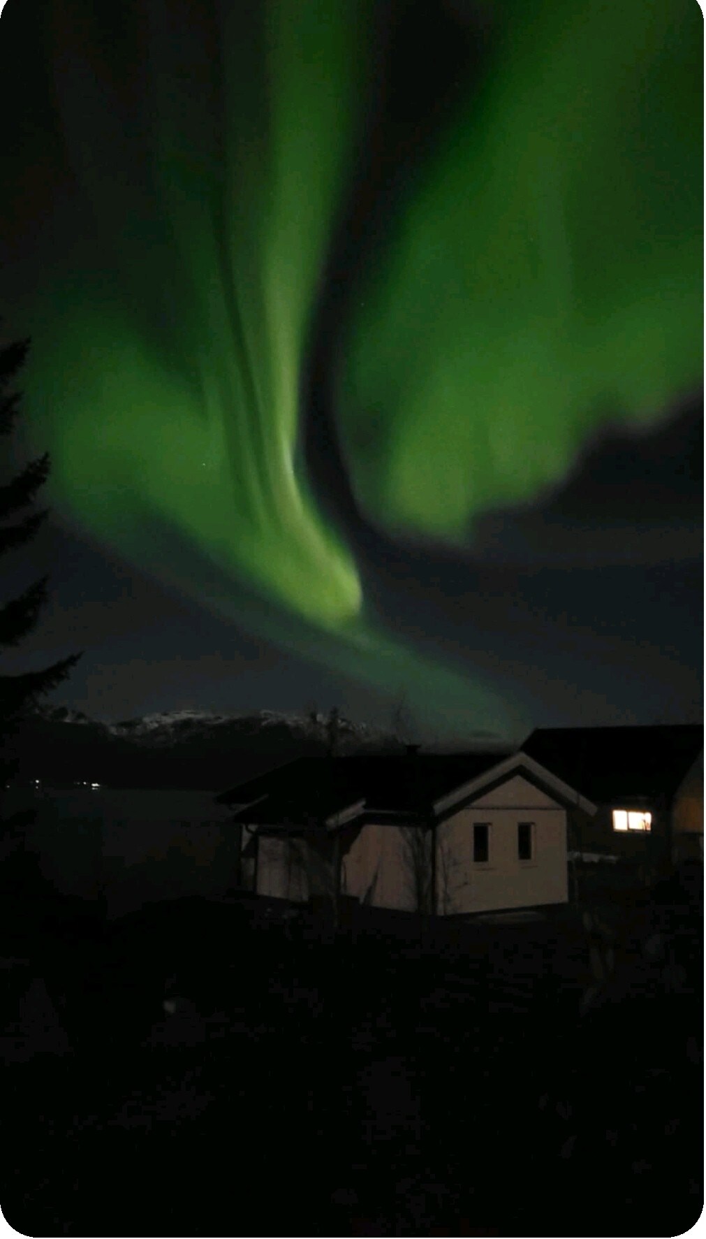 The northern lights illuminate the night sky with vibrant green hues, creating a dramatic display above the surrounding landscape. A cozy cabin is visible in the foreground, nestled in a serene setting with silhouetted trees. Mountain peaks can be faintly discerned in the distance.