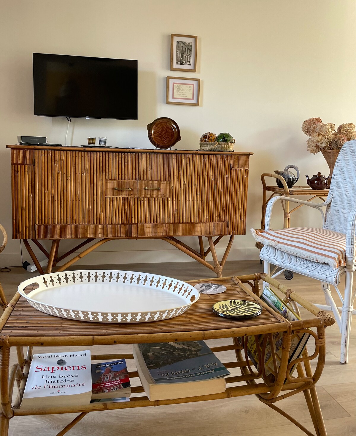 A welcoming living area features a wooden sideboard with decorative items and a small TV mounted on the wall. A bamboo coffee table holds a selection of books and a woven tray. Comfortable seating options are present, inviting relaxation.