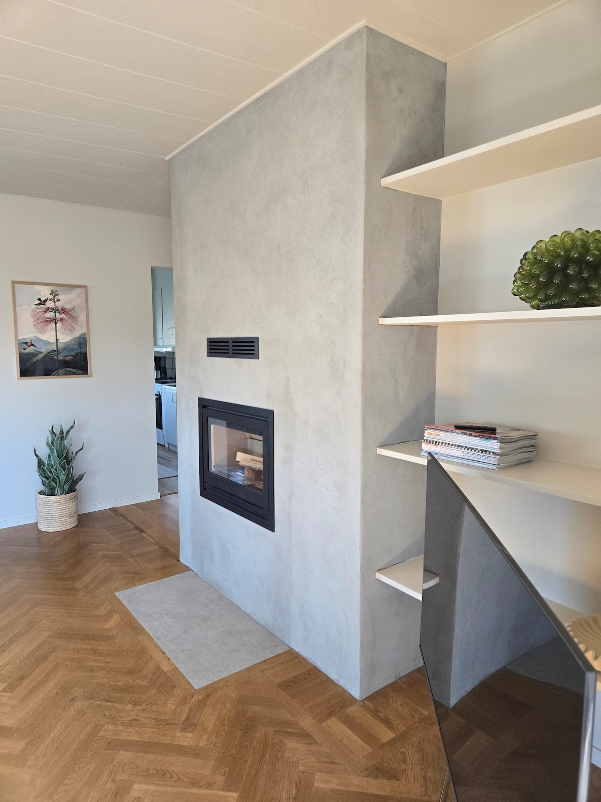 A modern fireplace is integrated into a smooth gray feature wall, providing a focal point for the room. Shelves are present on either side, displaying decorative items and books. Light hardwood flooring complements the neutral color palette of the space.
